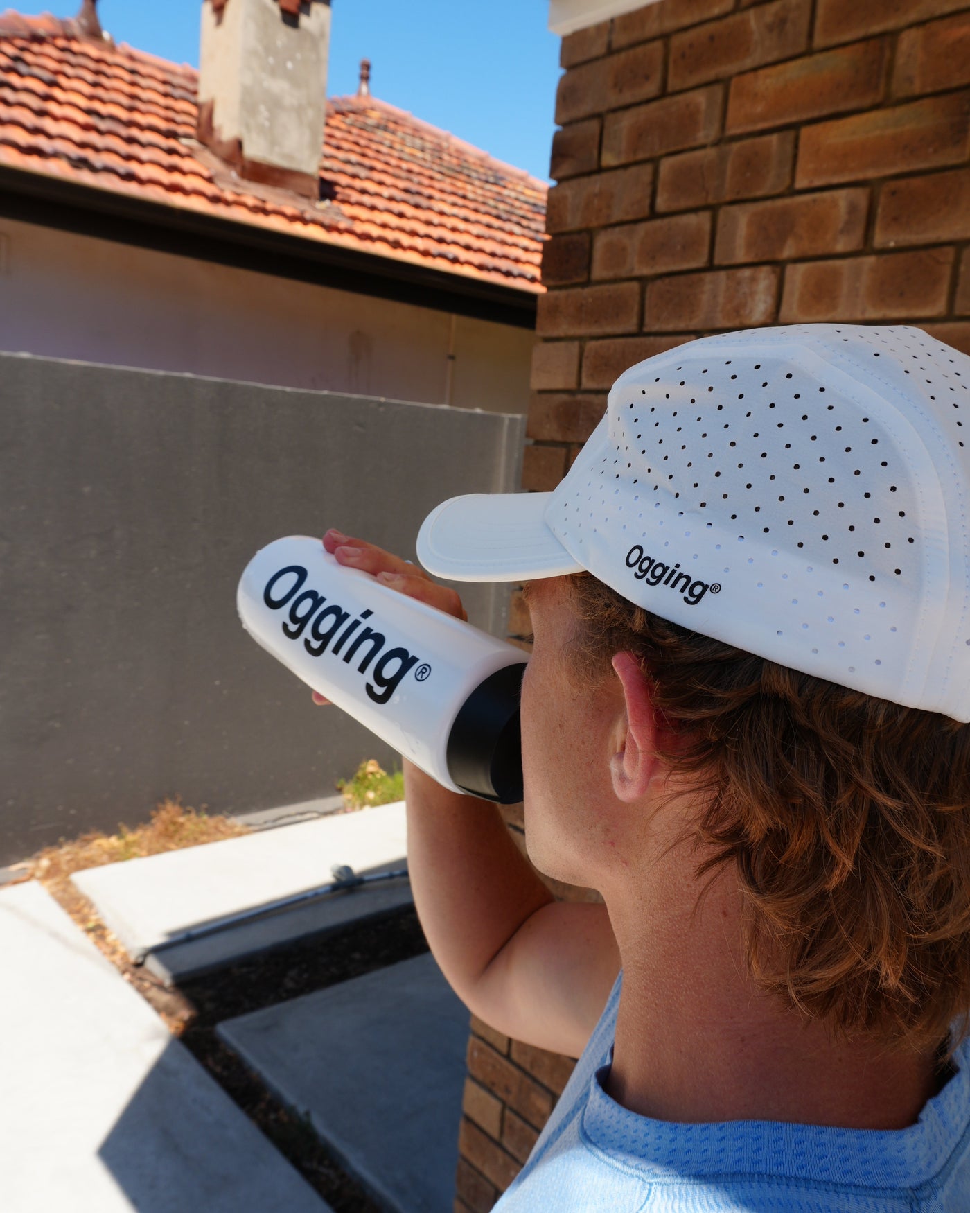 Person wearing a white cap and holding a black and white bottle with 'Ogging' branding, standing on a roof.