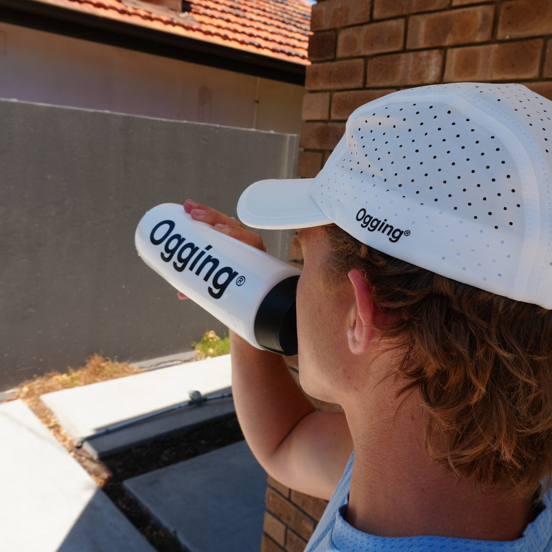Person wearing a white cap and holding a black and white bottle with 'Oggins' branding, standing on a roof.